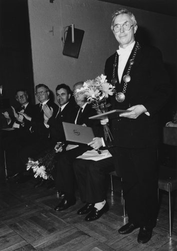 Mann mit Rektorkette hält Blumenstrauß und Urkunde in der Hand